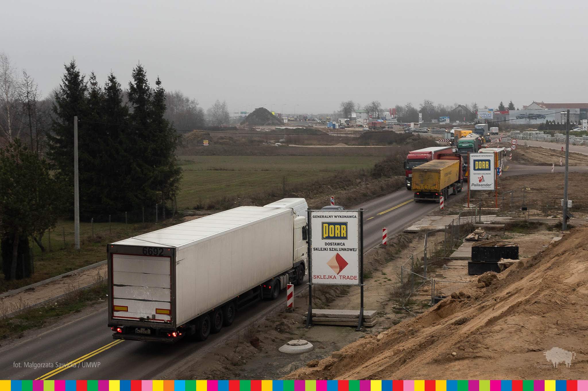 Zdjęcie przedstawia teren, na którym będą prowadzone prace budowlane - na dwupasmowej drodze widać ciężarówki jadące w dużym ścisku, po prawej stronie widać teren przygotowany do rozpoczęcia budowy z górą piasku. 