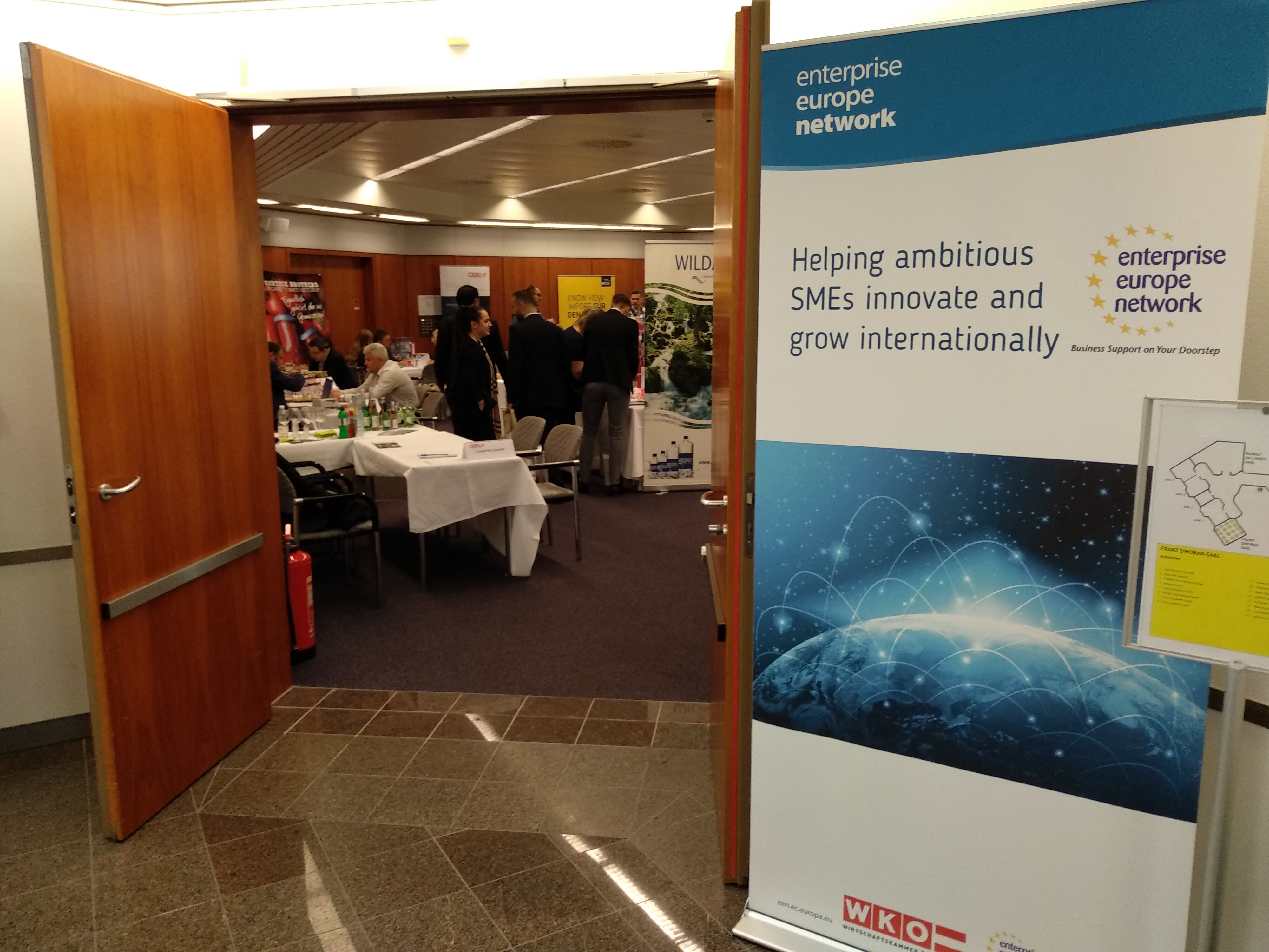 widok na korytarz i otwarte drzwi do sali, w której znajdują się ludzie. Obok drzwi widoczny roll-up z napisem Enterprise Europe Network