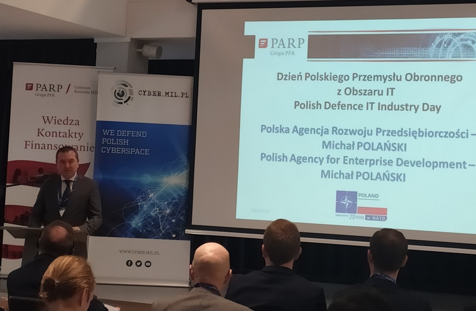 Na pierwszym planie po prawej stronie widać mężczyznę w średnim wieku, który stoi przy mównicy. Na...