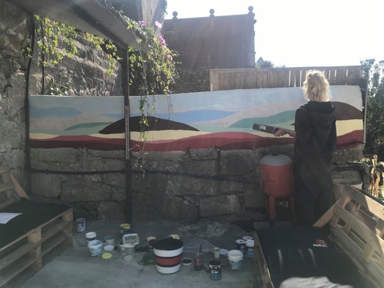 Młoda kobieta stoi przy murku na wysokość klatki piersiowej i maluje na nim abstrakcyjne pejzaże