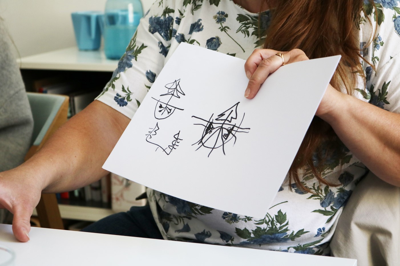 zbliżenie na ręce kobiety, która trzyma kartkę. Na białej kartce widać naniesiony czarny szkic, które wyglądem przypomina doniczkę wkopaną w ziemię