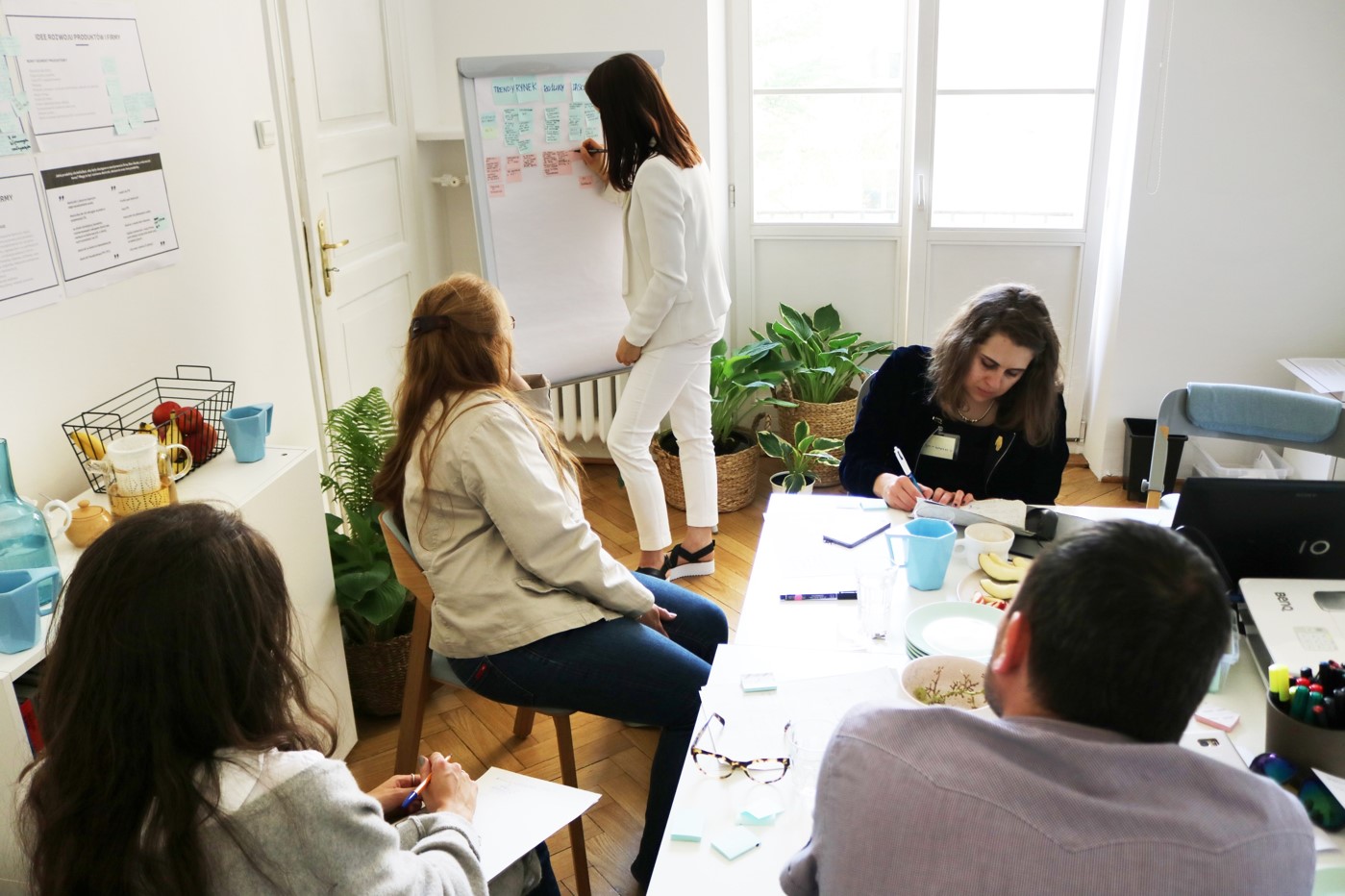 w niewielkim pomieszczeniu przy stole siedzą trzy kobiety w młodym lub średnim wieku oraz mężczyzna w młodym lub średnim wieku. Patrzą się w kierunku małej tablicy ustawionej przy ścianie. Na tablicy widać przyklejone małe karteczki w różnych kolorach, a obok tablicy stoi kobieta, która przykleja kolejną karteczkę