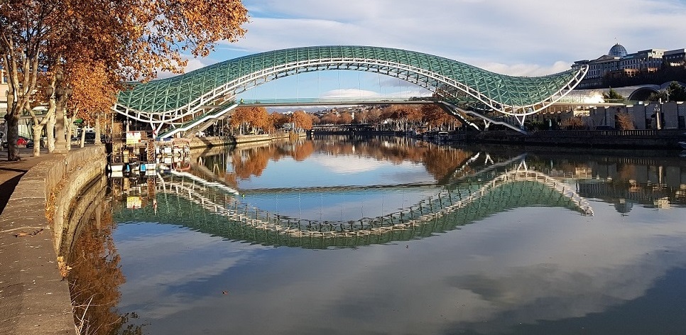 Widok na bulwary miejskie w Tbilisi, na pierwszym planie rzeka, w tle widać most. Po bokach zadrzewione bulwary rzeczne i zabudowania