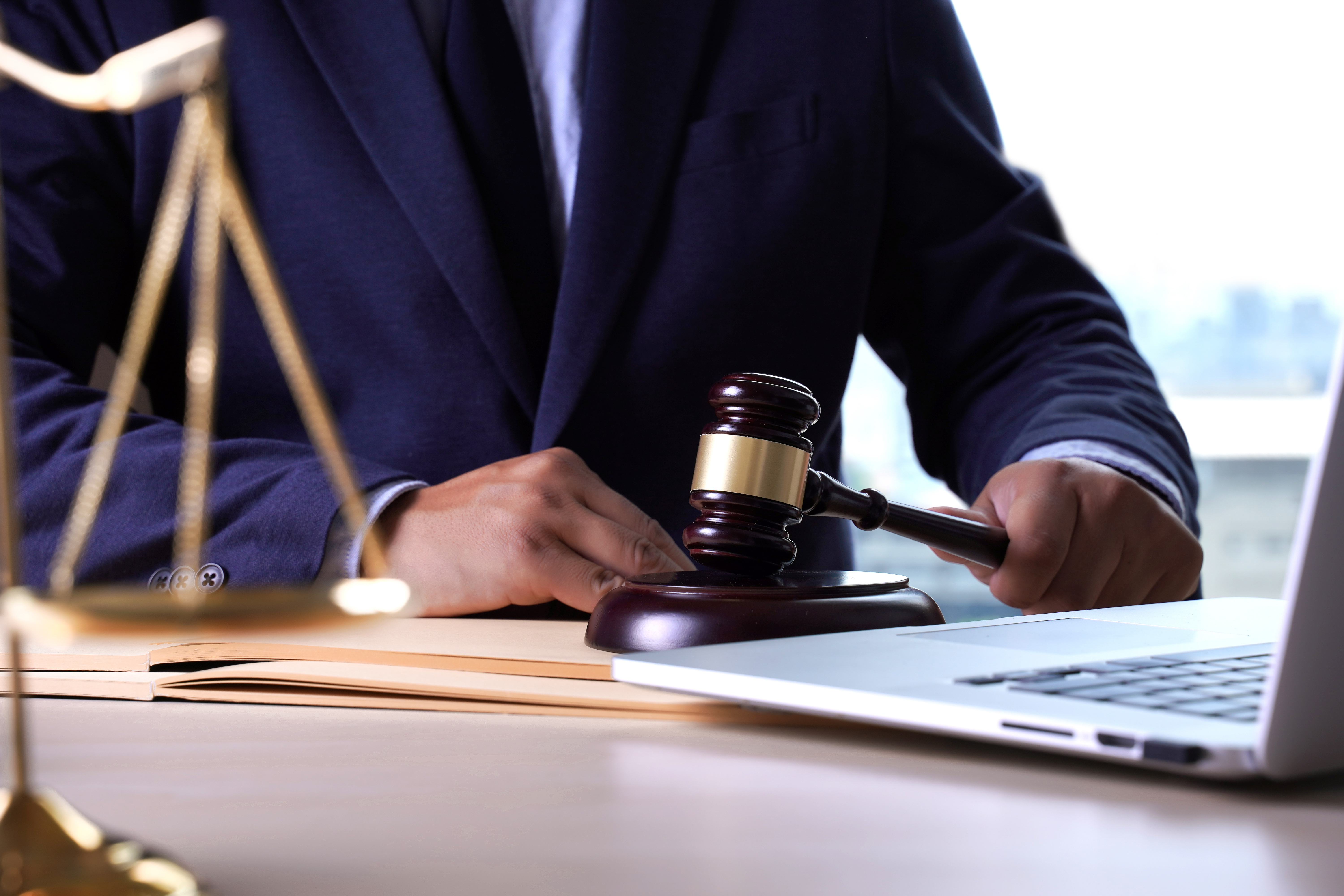 Zdjęcie przedstawia mężczyznę od szyi w dół siedzącego przy stole w granatowym garniturze. Mężczyzna ma dłonie o ciemniej karnacji, w jednej z nich trzyma drewniany młotek sędziowski i próbuje uderzyć o podstawkę na blacie.