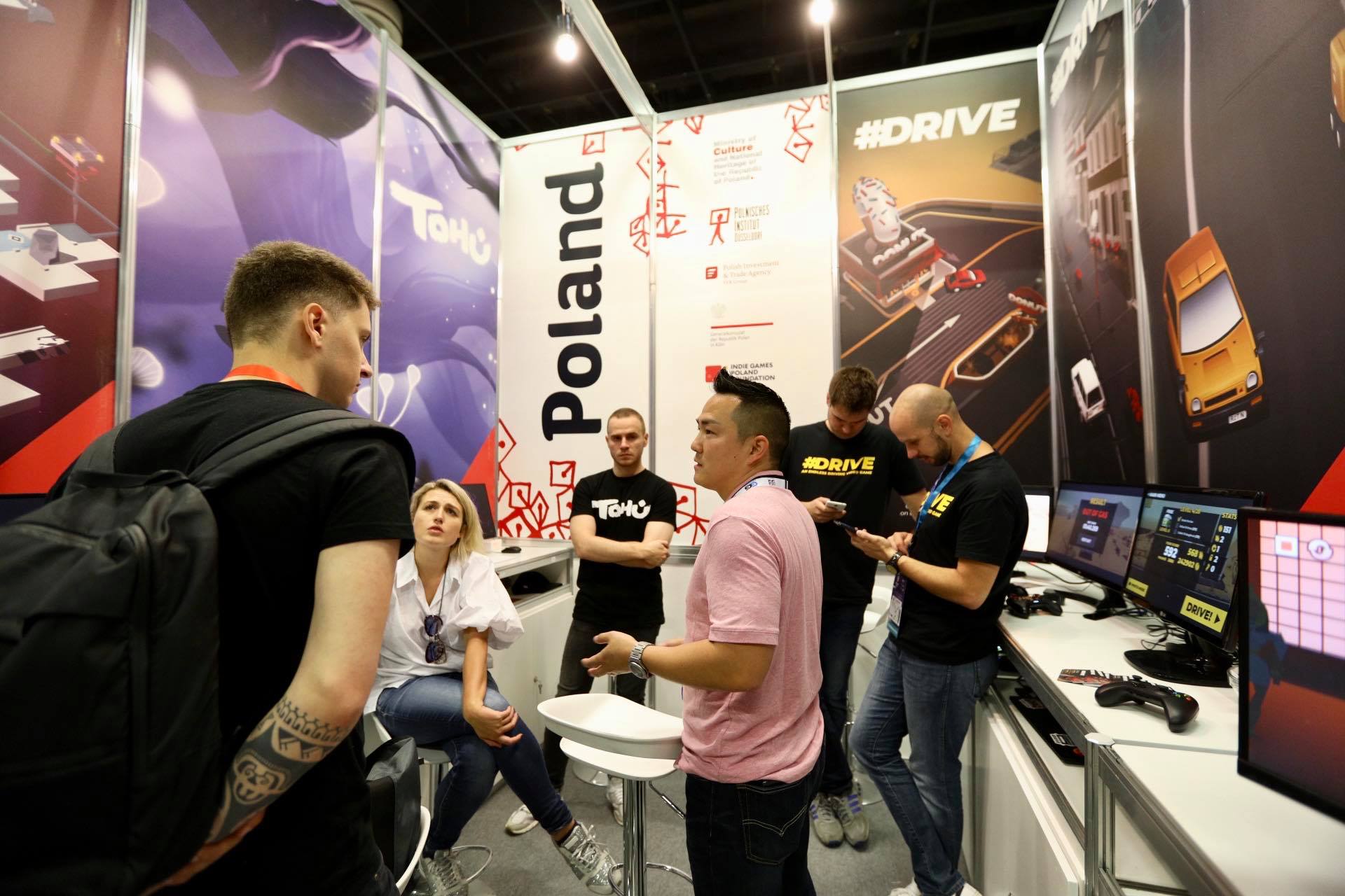 Na pierwszym planie trzy rozmawiające ze sobą osoby. W tle jedna osoba się im przygląda, a dwie patrzą na swoje smartfony. Po prawej stronie na stoliku widoczne trzy monitoryz włączonymi grami komputerowymi