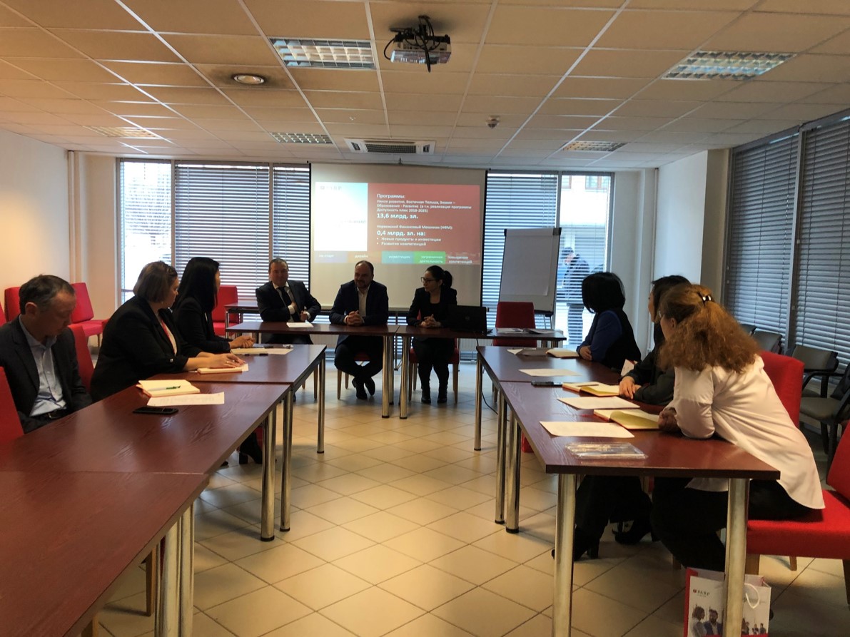 kobiety i mężczyźni w różnym wieku siedzą przy trzech stołach w sali. Na biurkach stołach leżą kartki. Stoły są ustawione względem siebie tworząc