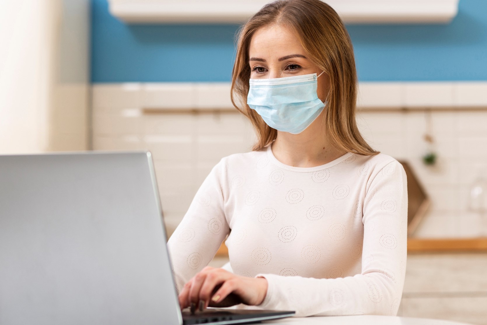 Młoda kobieta w maseczce hirurgicznej na twarzy pracuje przy komputerze, ubrana jest w jasną bluzkę
