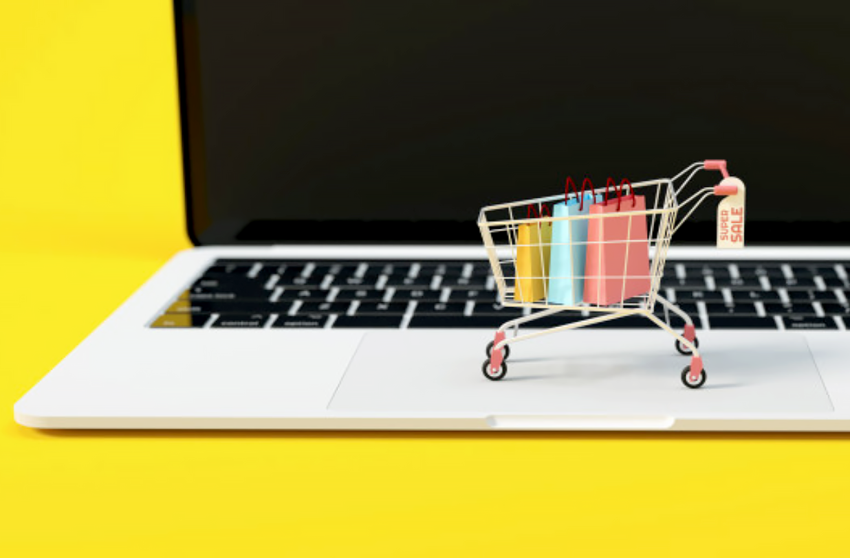 Na zdjęciu widać laptopa na żółtym tle. Na laptopie stoi miniaturowy koszyk zakupowy z trzema malutkimi torbami w środku.