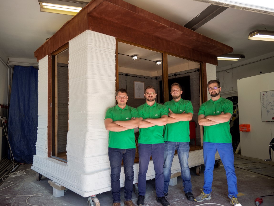 Czterech mężczyzn w zielonych koszulkach stoi przed małym betonowym domkiem umieszczonym w hali