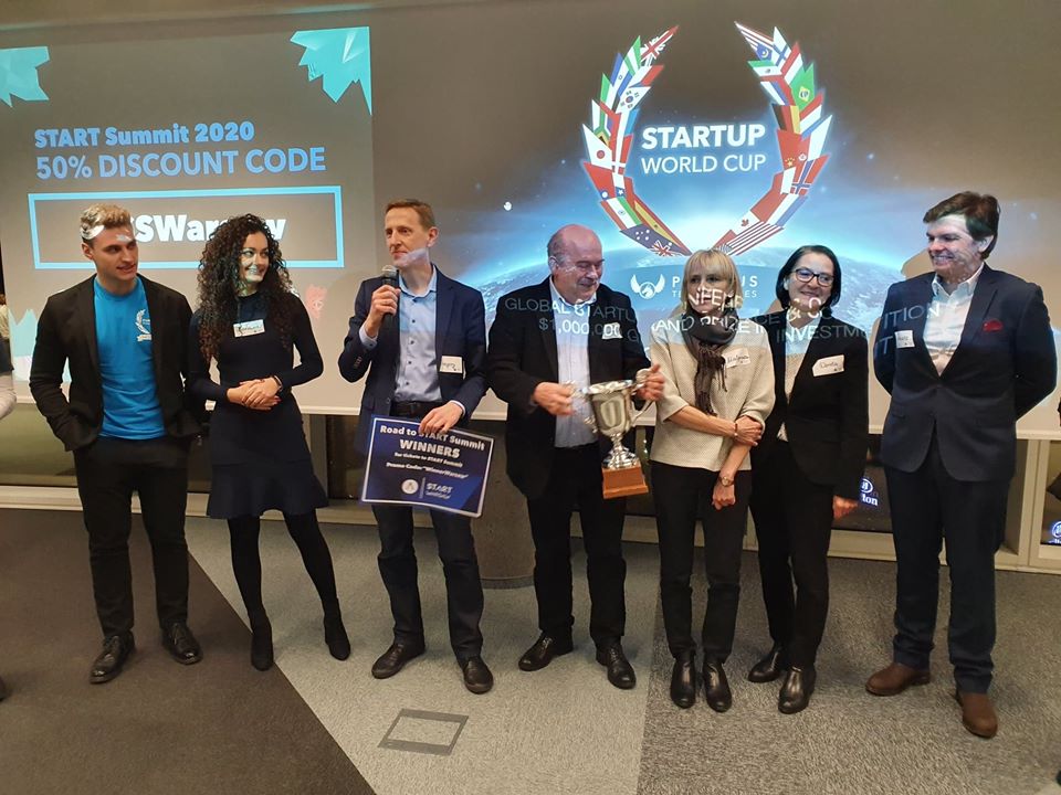 na zdjęciu widać siedem osób, trzy kobiet i czterech mężczyzn, w różnym wieku, stojących w sztucznie oświetlonym pomieszczeniu. Za nimi widać ekran z angielskojęzycznym napisem Startup World Cup. Jedna z osób trzyma w ręku puchar