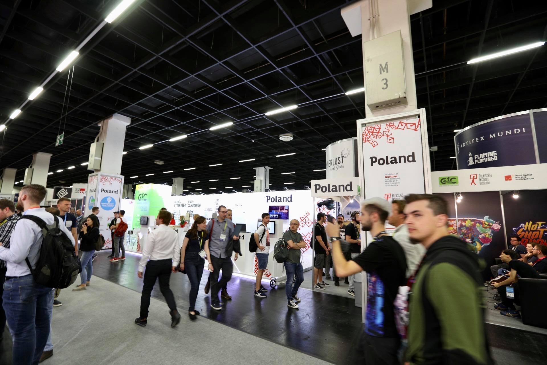 Ludzie przechodzący obok polskiego stoiska. Na stoisku widać białe ścianki i napisy