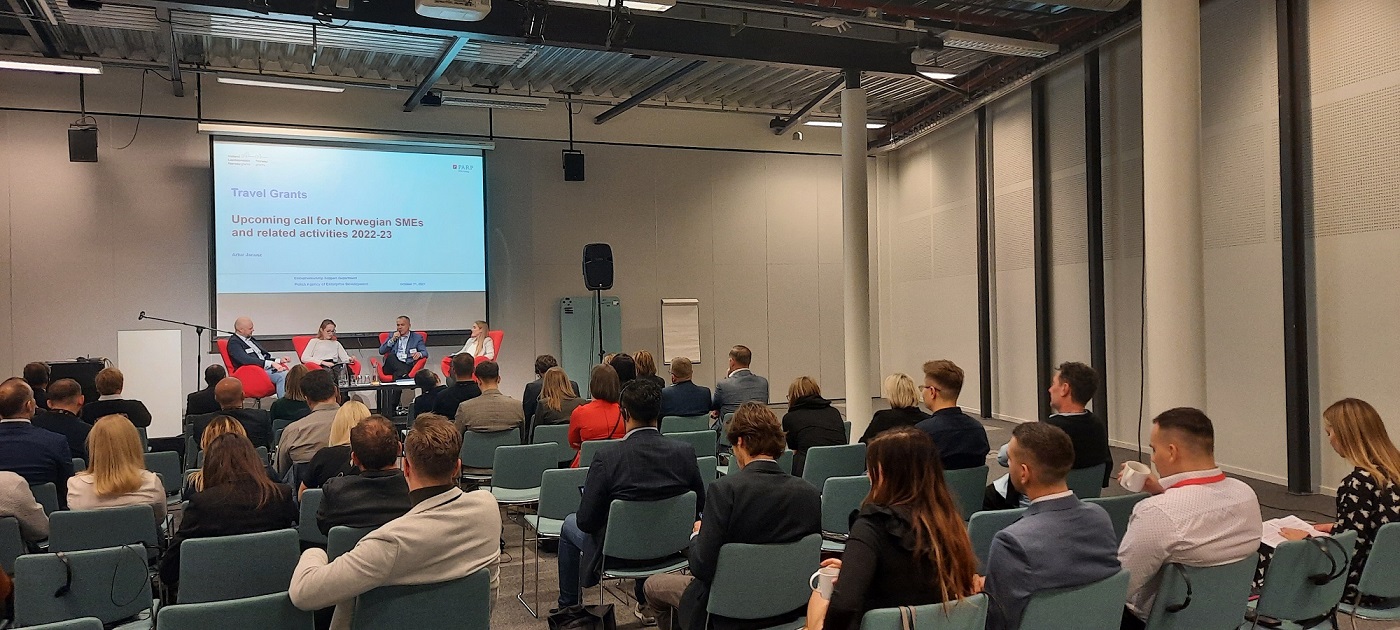 widok na ludzi zgromadzonych na sali konferencyjnej i siedzących na krzesłach na przeciwko sceny, na której siedzą cztery osoby, jedna z nich ma mikrofon w ręku