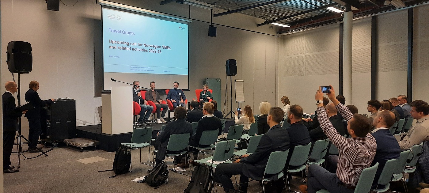 widok na salę konferencyjną z ludźmi siedzącymi na krzesłach i przysłuchującymi się wystąpieniu osoby stojącej na scenie