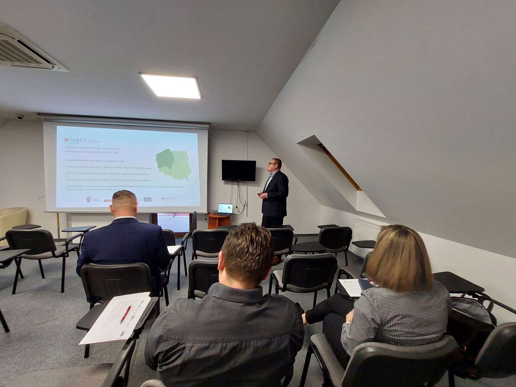 na zdjęciu prelegent dr Wojciech Bednarek  przedstawia ofertę GOZ (Gospodarka o obiegu zamkniętym) poprzez wyświetlenie na ekranie prezentacji, na pierwszym planie znajduje się publiczność