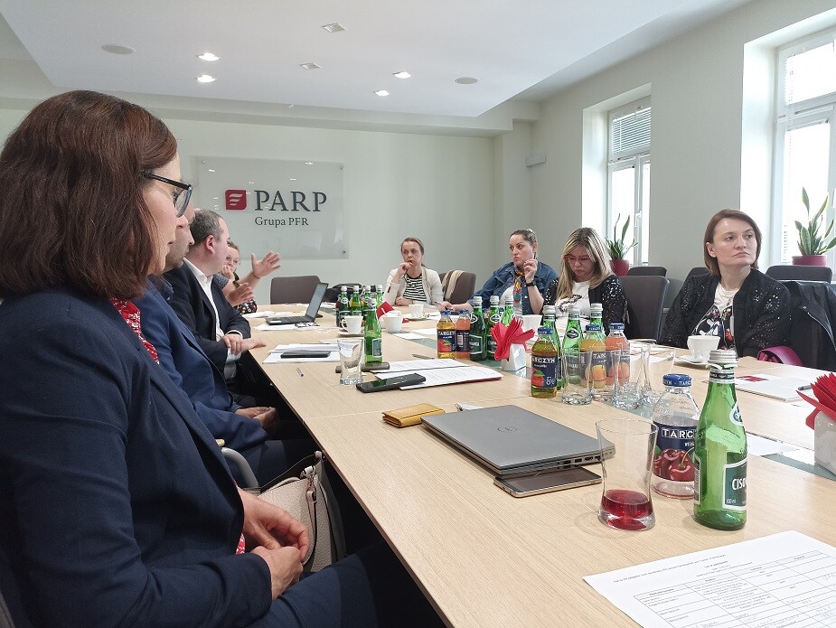 Ludzie siedzą przy stole konferencyjnym w podłużnej sali i rozmawiają 