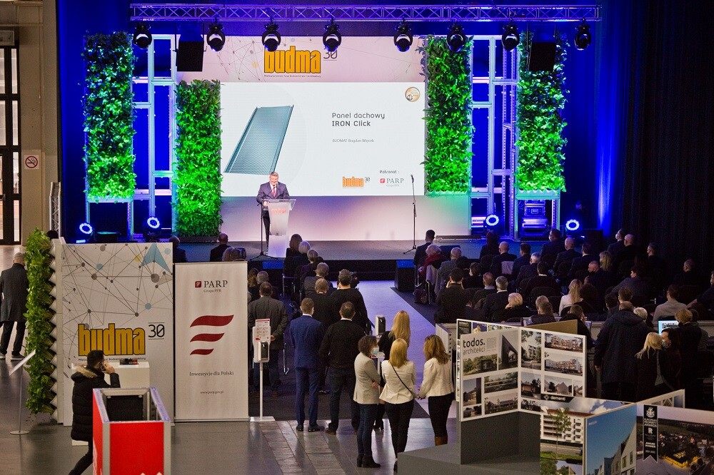 Widzimy uśmiechnięta kobiete w granatowej marynarce, stojącej za białym pulpitem z logo MTP, mówiącą coś do mikrofonu. Tło jest z zielonej roślinności