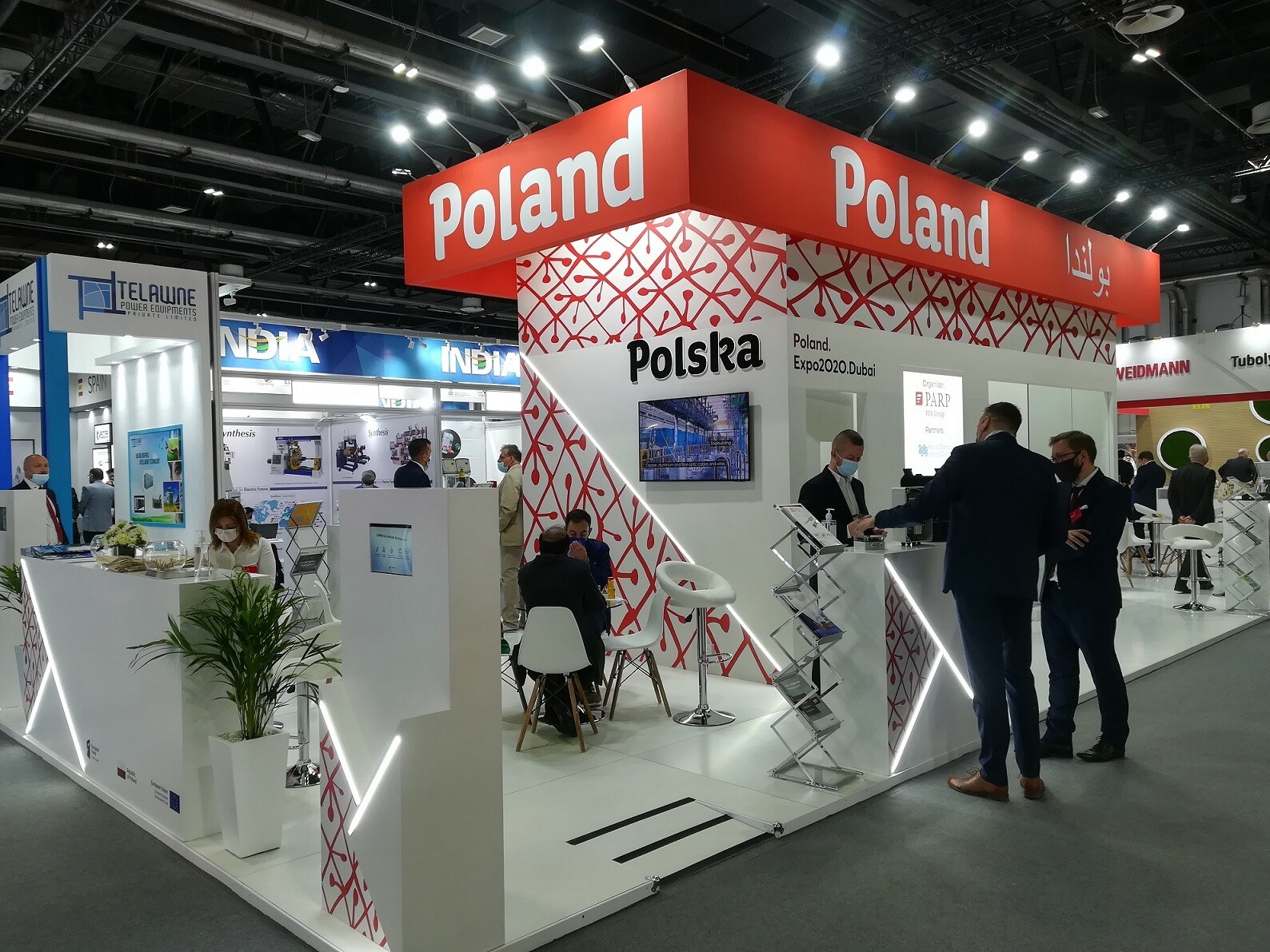 Widok na wnętrze hali wystawowej ze stoiskiem. Na górnej części kiosku widać napis "Poland". Przy stoisku stoją ludzie