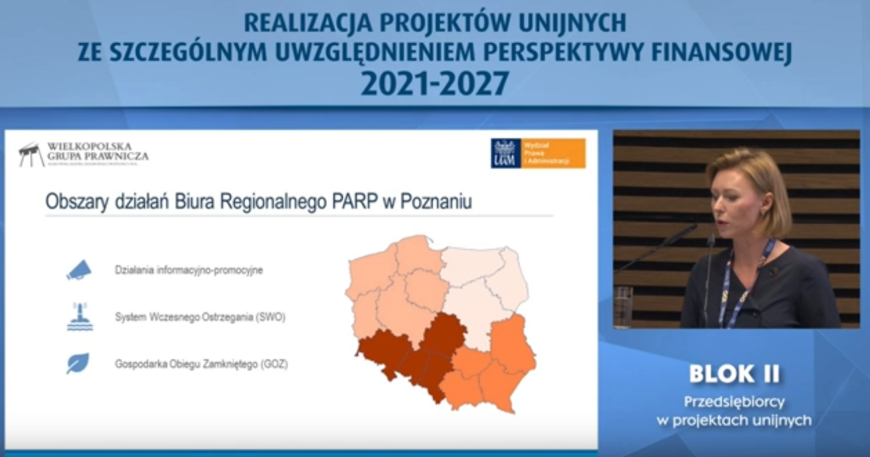 Zdjęcie przedstawia niebieski slajd podpisany –„realizacja projektów unijnych ze szczególnym uwzględnieniem perspektywy finansowej 2021-2027”.  Po lewej stronie widać na białym tle napisy „obszary działania Biura Regionalnego PARP w Poznaniu: działania informacyjno-promocyjne; system wczesnego ostrzegania SWO; gospodarka obiegu zamkniętego GOZ” oraz przedstawia mapkę Polski, a po prawej stronie widać kobietę mówiącą do mikrofonu. Kobieta ubrana jest na granatowo – pod zdjęciem widać napis „Blok II Przedsiębiorcy w projektach unijnych”.