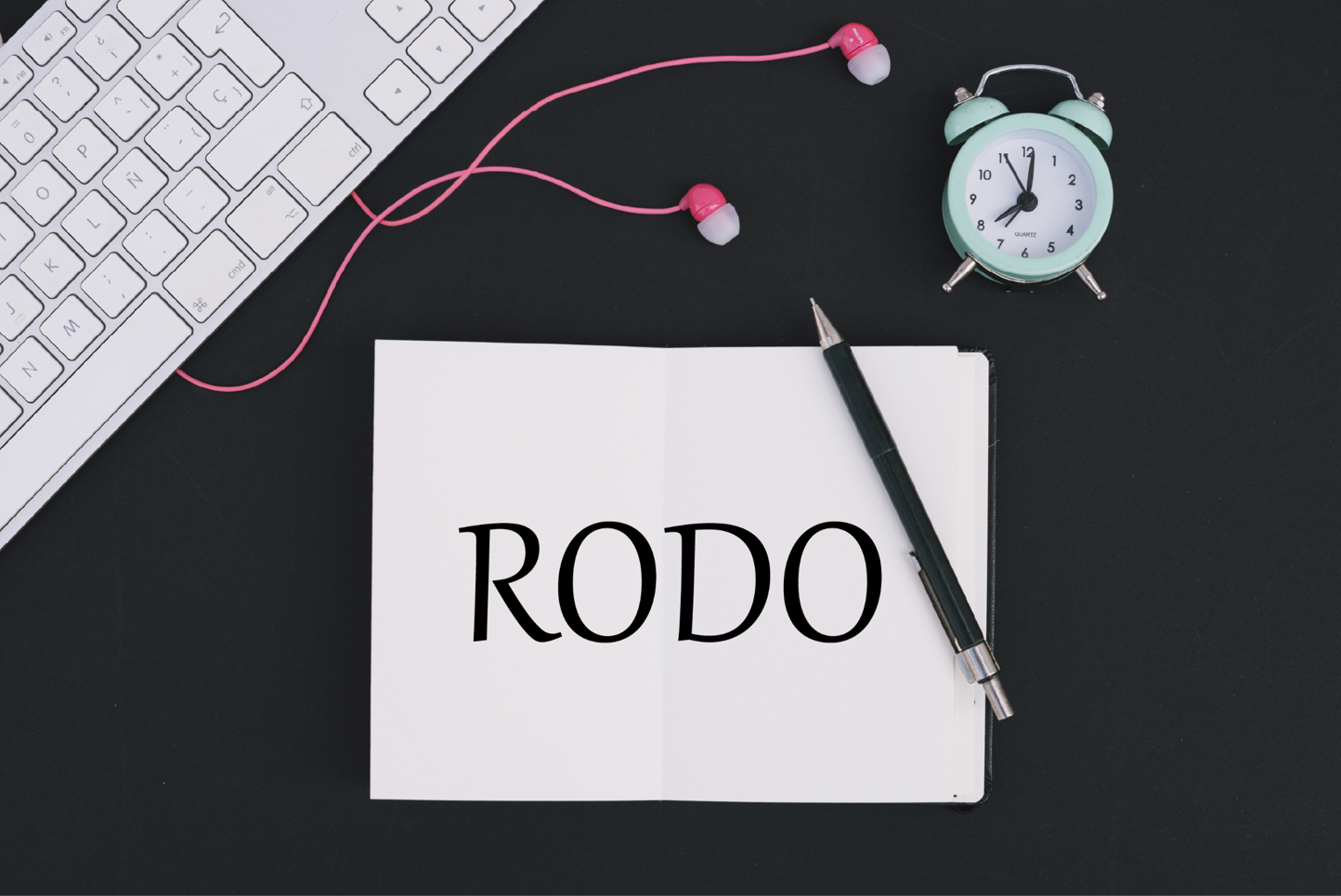 W centralnym punkcie zdjęcia znajduje się biała kartka z napisem: RODO. W lewym górnym rogu widoczny jest fragment klawiatury komputera. Na zdjęciu znajdują się również różowe słuchawki, budzik i długopis