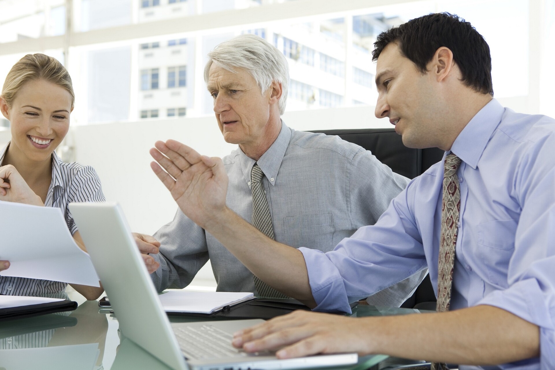 Widok na ludzi siedzących przy biurku, od lewej młoda kobieta, w  środku starszy mężczyzna o siwych włosach, po prawej młodszy mężczyzna w czarnych włosach. Ubrani w koszule z krawatami