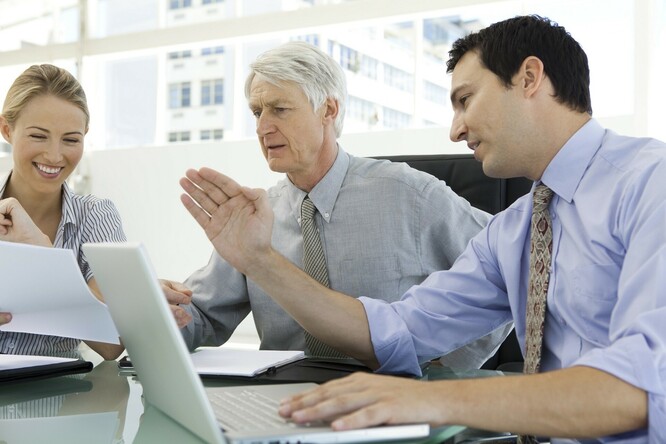 Widok na ludzi siedzących przy biurku, od lewej młoda kobieta, w  środku starszy mężczyzna o...