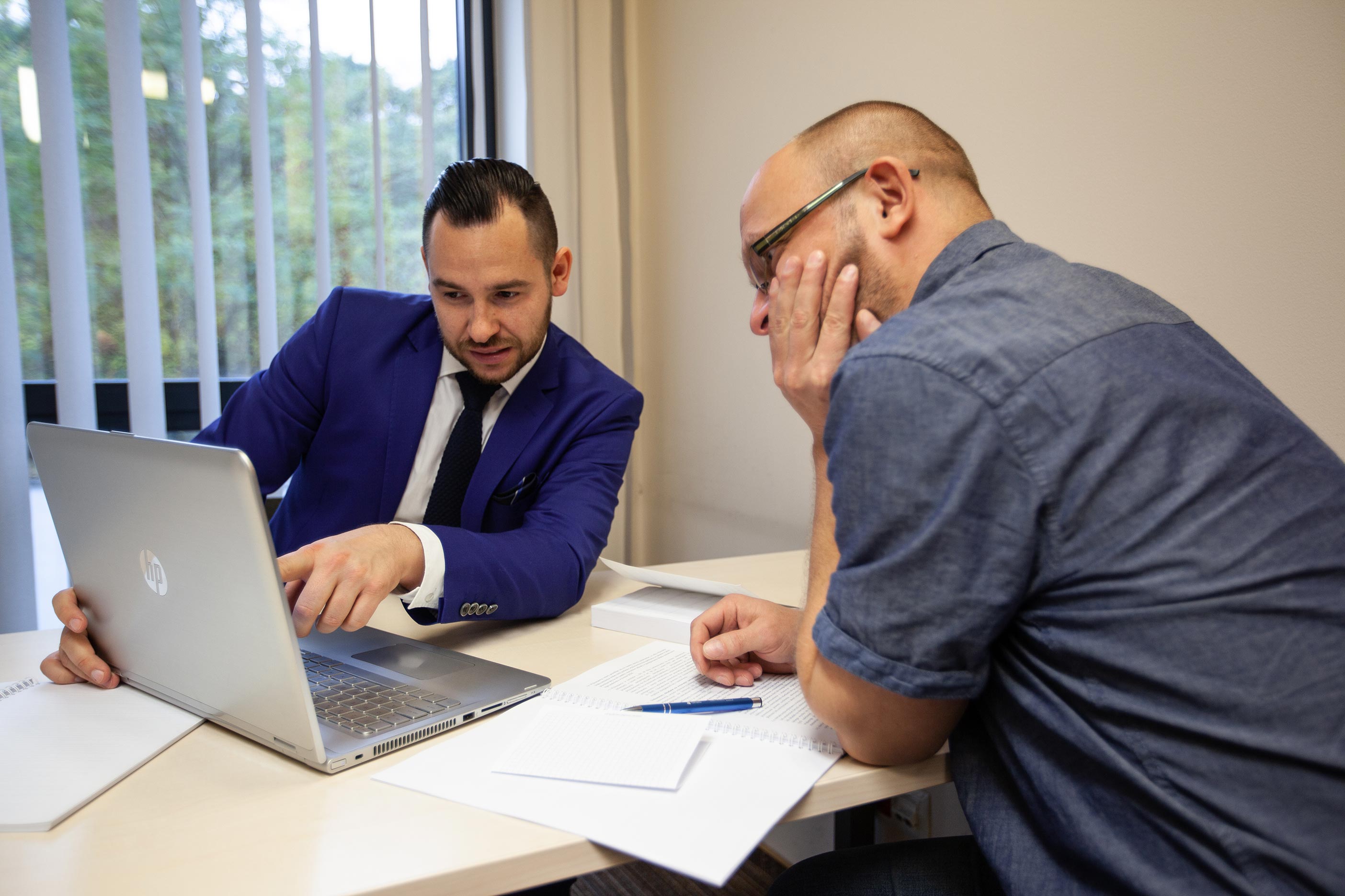 Widok dwóch mężczyzn przy biurku, jeden w garniturze pokazuje coś ręką na ekran komputera