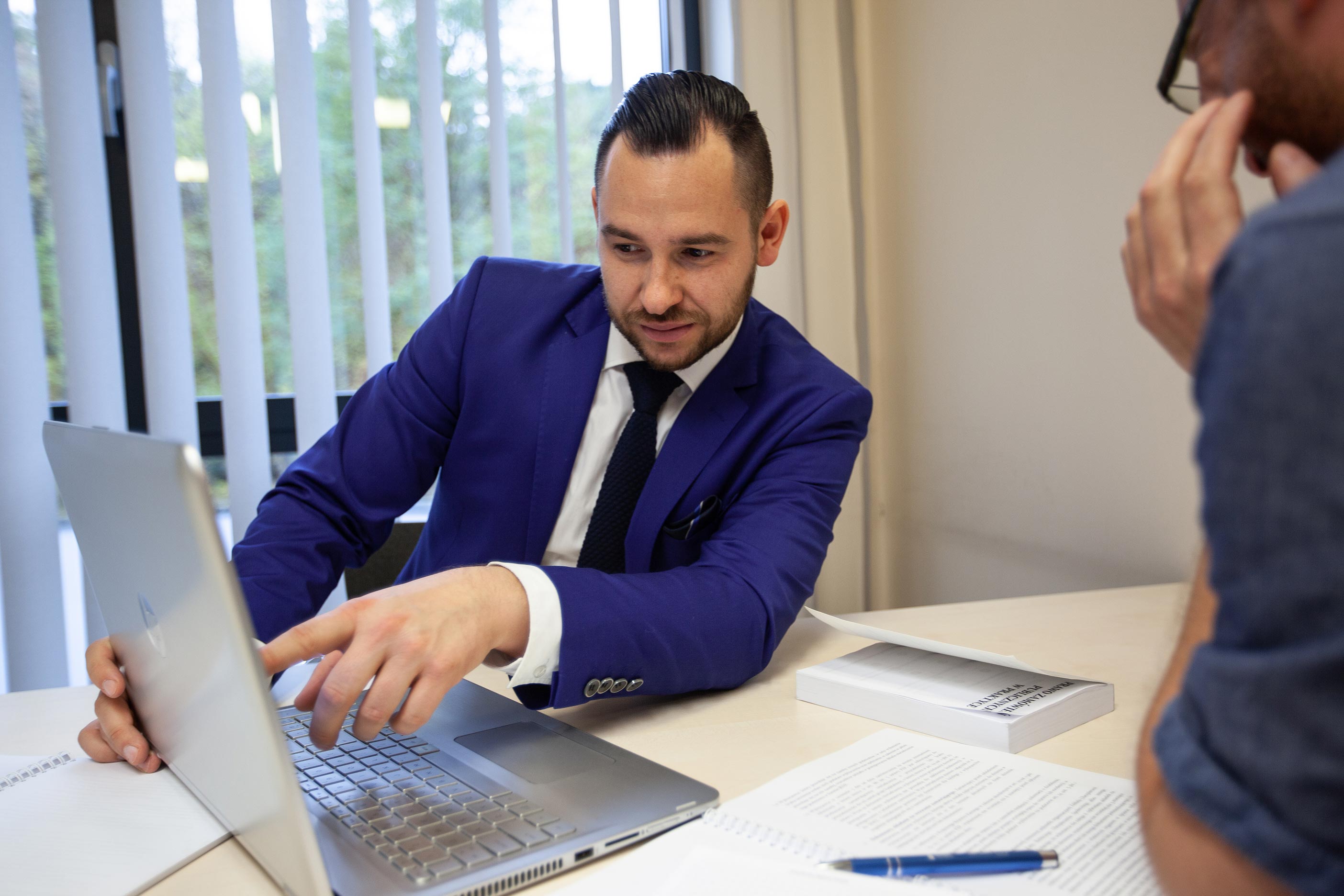 Widok dwóch mężczyzn przy biurku, przybliżenie osoby w garniturze, która pokazuje coś ręką na ekranie komputera