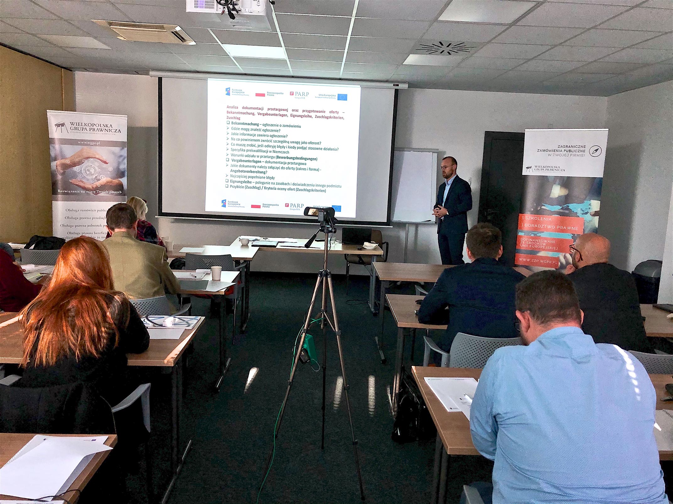 Sala szkoleniowa, uczestnicy siedzący przy stolikach, w tle prezentacja Zamówienia publiczne" i prelegent.